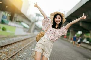 Jeune asiatique femme voyageur avec tissage panier content souriant à la recherche à une caméra à côté de train chemin de fer. périple voyage mode de vie, monde Voyage explorateur ou Asie été tourisme concept. photo