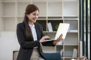 portrait, professionnel et sur de soi millénaire asiatique femme d'affaires ou femelle exécutif directeur dans formel costume et lunettes, debout, penché sur tableau, en portant portable et en utilisant portable ordinateur. photo