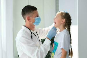 une médecin dans une médical masque examine le gorge de une enfant. une médecin examine une peu fille pendant un maladie. appel une médecin à maison. photo