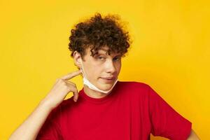 portrait de une Jeune frisé homme portant une rouge T-shirt médical masque sur le visage posant Jaune Contexte inchangé photo