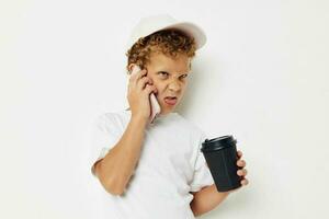 mignonne peu garçon dans une blanc T-shirt casquette avec une téléphone dans une verre avec une boisson lumière Contexte inchangé photo