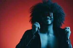 portrait de mode homme avec frisé cheveux sur rouge Contexte avec élégant lunettes, multiculturel, coloré lumière, noir cuir veste s'orienter, moderne concept. photo