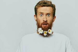 portrait de une marrant homme dans une blanc T-shirt avec fleurs marguerites dans le sien barbe sur une blanc isolé arrière-plan, copie lieu. vacances concept et toutes nos félicitations. photo