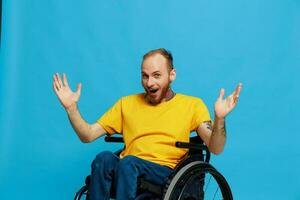 une homme est assis dans une fauteuil roulant prévenance dans une T-shirt sur une bleu Contexte dans le studio, élevé le sien mains en haut et ouvert le sien bouche dans surprendre, tatoué corps photo