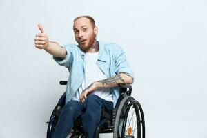 une homme dans une fauteuil roulant regards à le caméra spectacles une les pouces en haut, bonheur, avec tatouages sur le sien mains est assis sur une gris studio arrière-plan, santé concept homme avec handicapées photo