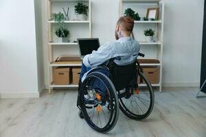une homme dans une fauteuil roulant pigiste travaux à une portable à maison, restriction de mouvement, vue de le dos, copie espace, l'intégration dans société, santé concept une la personne avec handicapées, une réel la personne photo