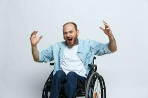 une homme dans une fauteuil roulant regards à le caméra colère et agression, avec tatouages sur le sien bras est assis sur une gris studio arrière-plan, santé concept homme avec handicapées photo