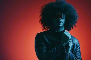 portrait de mode homme avec frisé cheveux sur rouge Contexte avec élégant lunettes, multiculturel, coloré lumière, noir cuir veste s'orienter, moderne concept. photo