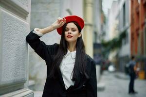 femme permanent près une mur dans le ville portant une élégant veste et rouge béret avec rouge lèvres, Voyage et loisirs, français style de robe, printemps. photo