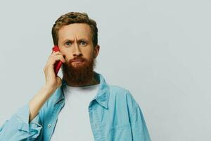 homme branché avec téléphone dans main blogueur sourire sur gris Contexte dans bleu chemise et blanc T-shirt, parlant sur le téléphone et selfies photo