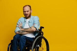 une homme dans une fauteuil roulant problèmes avec le musculo-squelettique système regards à le caméra est ne pas satisfait, avec tatouages sur le sien bras est assis sur une Jaune studio arrière-plan, santé concept homme avec handicapées photo