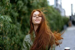 mode femme dans le ville à l'extérieur posant avec une sourire contre bâtiments et des arbres avec rouge longue en volant cheveux dans le venteux temps photo