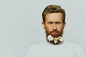 portrait de une marrant homme dans une blanc T-shirt avec fleurs marguerites dans le sien barbe sur une blanc isolé arrière-plan, copie lieu. vacances concept et toutes nos félicitations. photo