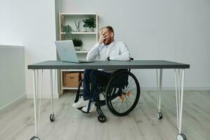 une homme fauteuil roulant, une homme d'affaire dans un Bureau à une ordinateur portable, fatigue et émotif Burnout à le fin de le travail jour, le concept de travail une la personne avec handicapées, liberté de social cadres photo