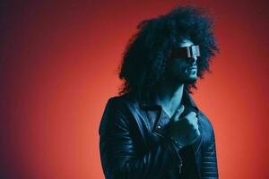 portrait de mode homme avec frisé cheveux sur rouge Contexte avec élégant lunettes, multiculturel, coloré lumière, noir cuir veste s'orienter, moderne concept. photo