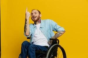 une homme dans une fauteuil roulant problèmes avec le musculo-squelettique système regards à le caméra spectacles une doigt sur, avec tatouages sur le sien mains est assis sur une Jaune studio arrière-plan, santé concept homme avec handicapées photo