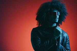 portrait de mode homme avec frisé cheveux sur rouge Contexte avec élégant lunettes, multiculturel, coloré lumière, noir cuir veste s'orienter, moderne concept. photo