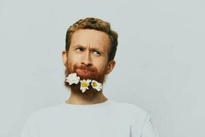 portrait de une marrant homme dans une blanc T-shirt avec fleurs marguerites dans le sien barbe sur une blanc isolé arrière-plan, copie lieu. vacances concept et toutes nos félicitations. photo