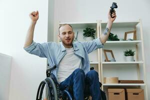 une homme dans une fauteuil roulant joueur pièces Jeux avec une manette dans le sien mains à maison, bonheur, la victoire, mains en haut, copie espace, avec tatouages sur le sien mains, santé concept homme avec handicapées, réel la personne photo