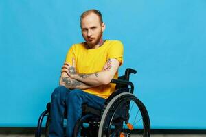 une homme dans une fauteuil roulant dans une T-shirt avec tatouages sur le sien bras est assis sur une bleu studio arrière-plan, une plein vie, une réel la personne photo