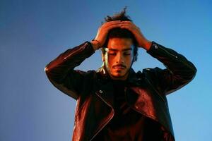 portrait de une élégant homme avec frisé cheveux sur une bleu Contexte multinational, coloré lumière, noir cuir veste s'orienter, moderne concept. photo