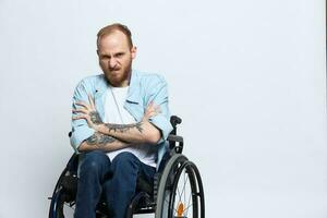 une homme dans une fauteuil roulant regards à le caméra colère et agression, avec tatouages sur le sien bras est assis sur une gris studio arrière-plan, santé concept homme avec handicapées photo