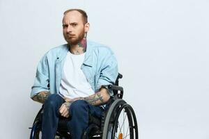 une homme dans une fauteuil roulant regards à le caméra, avec tatouages sur le sien bras est assis sur une gris studio arrière-plan, le concept de santé est une la personne avec handicapées photo