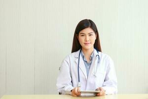 asiatique femelle médecin séance dans Bureau en portant une tablette à la recherche à traitement information. concept de médical prestations de service dans hôpitaux. en ligne médecin photo