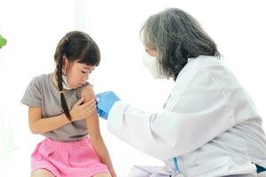 médecins injecter antiviral vaccin à mignonne peu asiatique fille avec nattes. asseoir sur le blanc lit. concept de vaccination pour les enfants à prévenir corona virus. blanc Contexte photo