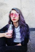 heureuse belle adolescente avec des lunettes de soleil roses boit et apprécie une boisson rose assise sur le sol urbain