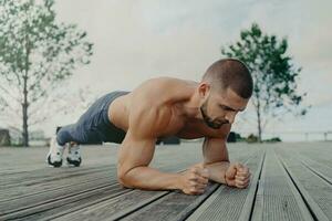 fort athlète maintient planche pose, donner la priorité aptitude et santé. concentré homme des exercices en plein air, soulignant abdominal force. bodybuilder se concentre sur coeur entraînement. photo