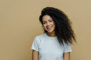 magnifique femme avec crépu cheveux, doux sourire, en bonne santé peau, décontractée blanc T-shirt, converser, beige Contexte. personnes, émotions. photo