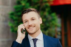 réussi Masculin ouvrier discute affaires sur téléphone, formel tenue le déjeuner Pause à café, positif sourire pose dehors. photo