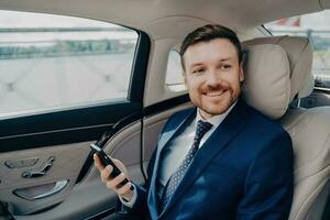 de bonne humeur exécutif dans élégant smoking chèques Stock des prix sur téléphone portable, monte à financier réunion dans luxe voiture avec conducteur. photo