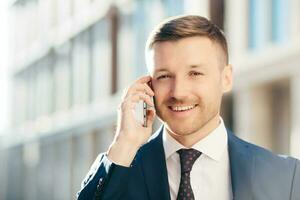 photo en extérieur d'un homme d'affaires mal rasé prospère et prospère porte des vêtements formels, a une conversation agréable avec un partenaire commercial via un téléphone portable, discute d'une future réunion, pose sur un arrière-plan flou