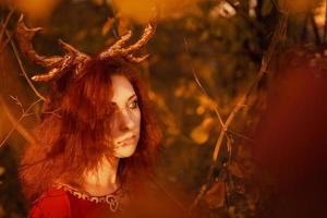 femme en longue robe rouge avec des cornes de cerf dans la forêt d'automne.