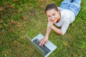 Jeune femme mensonge sur vert herbe pelouse dans ville parc travail sur portable PC ordinateur. free-lance affaires concept photo