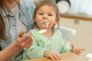 content famille à maison. mère alimentation sa bébé fille de cuillère dans cuisine. peu bambin enfant avec désordonné marrant visage mange en bonne santé nourriture à maison. Jeune femme maman donnant nourriture à enfant fille. photo