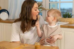 content famille à maison. mère alimentation bébé dans cuisine. peu garçon avec désordonné marrant visage mange en bonne santé aliments. enfant apprend manger par lui-même en portant cuillère. femme maman donnant nourriture à enfant fils. soi alimentation. photo