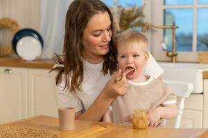 content famille à maison. mère alimentation sa bébé garçon de cuillère dans cuisine. peu bambin enfant avec désordonné marrant visage mange en bonne santé nourriture à maison. Jeune femme maman donnant nourriture à enfant fils. photo