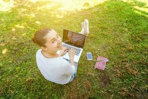 jeune femme assise sur une pelouse d'herbe verte dans le parc de la ville travaillant sur un ordinateur portable. concept d'entreprise indépendant photo