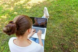 jeune femme assise sur une pelouse d'herbe verte dans le parc de la ville travaillant sur un ordinateur portable. concept d'entreprise indépendant photo