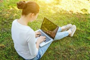 jeune femme assise sur une pelouse d'herbe verte dans le parc de la ville travaillant sur un ordinateur portable. concept d'entreprise indépendant photo