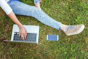 jambes de femme sur la pelouse d'herbe verte dans le parc de la ville, mains travaillant sur un ordinateur portable. concept d'entreprise indépendant photo