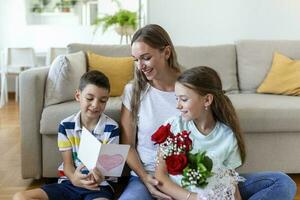 Jeune mère avec une bouquet de des roses des rires, étreindre sa fils, et de bonne humeur fille avec une carte et des roses félicite maman pendant vacances fête à maison. les mères journée photo