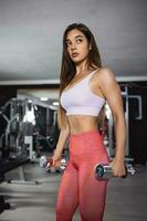 en forme fort Jeune femme levage poids travail en dehors avec haltères permanent avec sa côté à le caméra fléchissement sa bras dans le salle de sport. photo