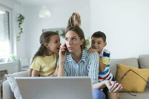 stressé femme avec des gamins travail de maison.fatigué Jeune mère séance sur canapé et travail avec portable et les documents tandis que peu des gamins ayant amusement et fabrication bruit photo