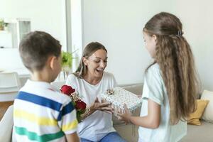 content les enfants donnant cadeau un fleurs à mère. content les mères journée. les enfants garçon et fille féliciter souriant mère, donner sa fleurs bouquet de des roses et une cadeau boîte pendant vacances fête photo