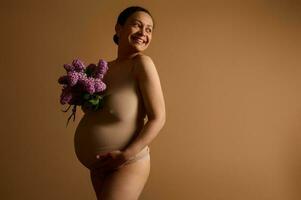 magnifique femme enceinte mère sourit à la recherche de côté une copie espace, posant dans beige lingerie avec lilas sur studio Contexte photo