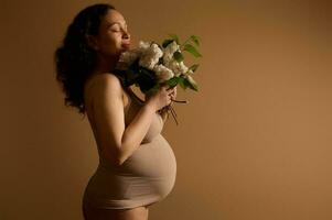 attrayant ethnique Enceinte femme, femme enceinte mère avec une bouquet de lilas, posant dans beige lingerie. studio portrait photo
